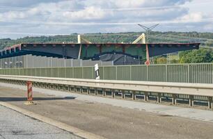 A view of a large, modern building with a distinctive roof design, partially obscured by a fence. The scene is set alongside a highway, with construction barriers visible in the foreground. photo