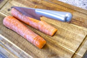 A close-up view of two fresh carrots on a wooden cutting board, with a sharp knife positioned above them, ready for slicing. The vibrant orange color of the carrots contrasts with the brown wood. photo