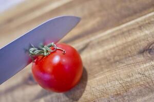 A close-up of a shiny red tomato on a wooden cutting board, with a sharp knife poised above it, ready to slice. photo