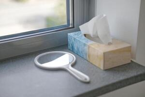 A close-up view of a window sill featuring a round hand mirror mirror and a decorative tissue box. The mirror has a white handle, and the tissue box is designed with a blue and beige pattern photo
