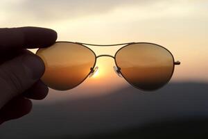 A person holding up a pair of sunglasses with the sun setting in the background photo