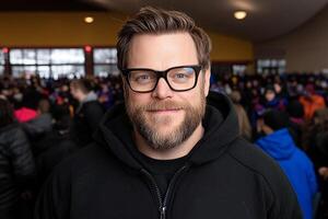 A man with glasses and a black jacket standing in front of a crowd photo