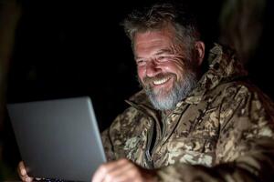 A man in camouflage is smiling while using a laptop photo