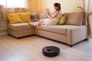 A robot vacuum cleaner washes the floor while a young woman reads a book sitting on the sofa. Smart home, automation and modern technologies in everyday photo