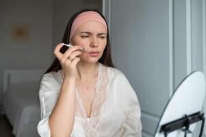 A middle-aged woman with already visible wrinkles in a silk robe gives herself beauty injections with a worried expression while looking in the mirror photo