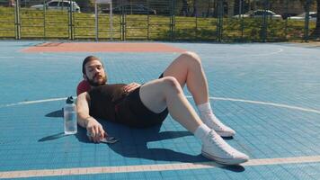 Tired man in sportswear after playing basketball game lying down on ball relaxing using smartphone video