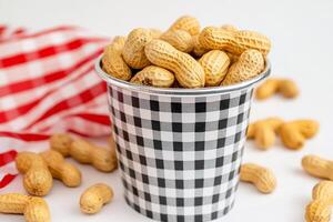 A cup filled with peanuts surrounded by scattered nuts and a checkered cloth. photo