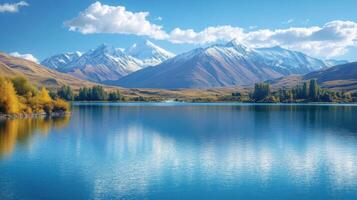 Calm lake water reflects blue sky and mountains. Autumn trees show gold colors on shore. Peaceful nature scenery with mountains. photo