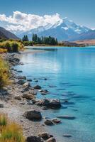 Lake view. Blue water reflects sky. Snow mountain and trees in distance. Rocks are on the shore. Peaceful scenery. Calm scene. photo