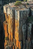 Tall stone cliff face. Golden light touches rocky canyon wall. Small plants grow at the top of stone formation. Rock face edge. photo