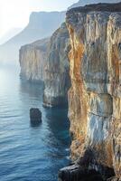 Rocky cliffs edge meets blue sea. Beautiful light shines on stone face. Calm ocean water washes against rocks. Distant mountains fade into horizon. photo