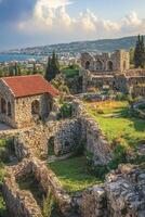 Old stone buildings. Ruins sit on hill. Green grass and stone. Town near blue water. White clouds in sky above. photo