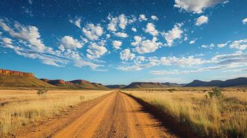 Dirt Road Landscape Under Blue Sky. Dry grass field with trail. Mountains in distance with clouds and stars above. photo