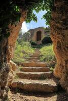 Stone steps lead to old ruin. Cave entrance frames a weathered building. Greenery surrounds the path. Bright sky shines above. A peaceful outdoor scene. photo
