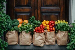 Fresh produce in brown paper bags sits by a dark wood door with green leafy plants on gray concrete. Tomatoes oranges and lettuce. photo