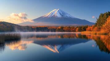 Snow capped mountain and autumn trees reflect on calm lake water under a blue sky with soft morning light and light mist. photo