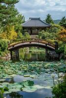 Japanese garden with wooden bridge crossing water lily pond leading to traditional building surrounded by green trees and hills under cloudy sky. photo