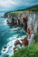 Rocky cliffs with cave openings stand above ocean. Waves crash on rocks below. Green grass covers the cliff tops. Mountains in the background under cloudy sky. photo