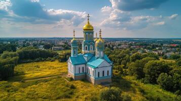 Aerial view of church with golden domes and blue roof tiles. Building surrounded by green trees and yellow meadow under a cloudy sky. photo