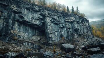 Steep stone wall with a cave opening and scattered rocks below. Cloudy sky above. Autumn foliage on trees atop the wall and nearby hillside. photo