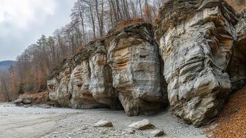 Layered rock cliffs stand on a rocky shore under a forest with mostly bare trees on an overcast day with grey skies. photo