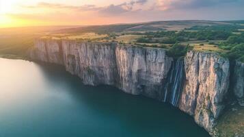 Scenic landscape with tall cliffs overlooking calm water. A waterfall cascades down the cliffside under a warm colorful sky. Green vegetation covers the cliff tops. photo