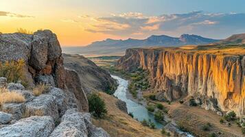 River flows through a canyon. Cliffs with yellow grass are lit by setting sun. Mountains are in distance with blue sky and light clouds. photo
