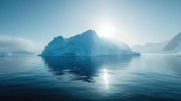 Large iceberg floats in calm arctic ocean water. Bright sun shines behind glacial ice. Mountain peaks visible under a clear blue sky. photo