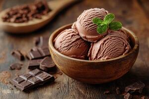 Chocolate ice cream scoops in wooden bowl. Mint leaf on top. Dark chocolate pieces and wooden spoon with chocolate shavings on rustic table. photo