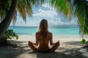 Woman sits meditating on sandy beach under palm trees facing calm turquoise ocean. Sunlight on ocean surface and cloudy blue sky backdrop. photo