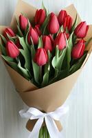 Bouquet of red tulip flowers wrapped in brown paper with white ribbon. Fresh spring blooms with green leaves against light backdrop. photo