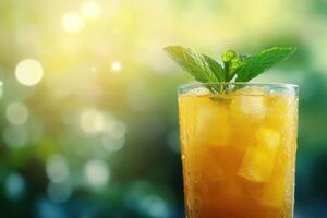Iced drink with mint leaves in a glass. Orange liquid with ice cubes. Blurred green nature background with sunlight. Refreshing cold beverage. photo