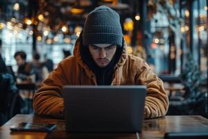 Man in warm clothes works on laptop at wooden table in cafe. Lights shine above. Others sit in background. photo