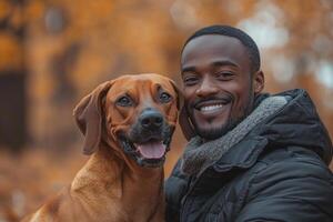 Smiling man poses with dog. Brown dog shows tongue. Fall backdrop with yellow leaves. Man wears black jacket and scarf. photo