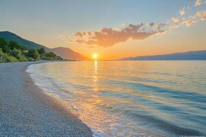 Calm sea waters gently wash over a pebble beach under a peaceful sunrise with distant mountains and scattered clouds above a golden horizon. photo