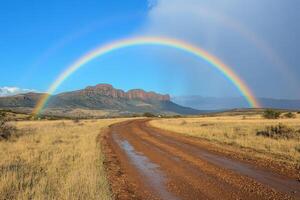Landscape with a mountain and field under a blue sky displaying a full beautiful rainbow above a muddy road through the grass. photo