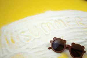 White sand spelling SUMMER on a bright yellow background, with sunglasses placed beneath the text. vacation vibe. photo