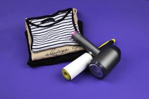 Clothes roller and lint machine are lying on clothes and purple background. In the background are several T-shirts of different colors. photo