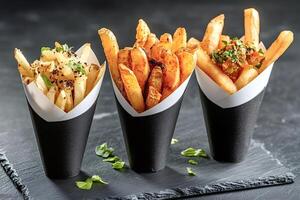 French fries with different toppings resting in black paper cones on slate plate photo