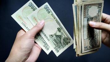 A person's hand is seen counting old rupiah banknotes of Rp10 to Rp500, isolated on a black background. Close-up view photo