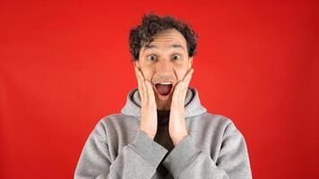 large close up portrait of young curly haired man, joyfully surprised, holding his face with his hands and mouth open, pronouncing word wow on studio red background. Concept - discounts, Black Friday photo