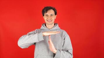 young adult curly haired brunette man holds his hands in front of shows sign timeout gesture. smiling, wearing grey hoodie. isolated on red studio background. Concept - pause, break, rest, vacation photo
