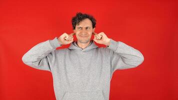 man in gray hoodie covers ears with fingers, does not want to listen to screams, loud music, avoids stress. Isolated on red background studio shot. concept - mental health, refusal to hear information photo