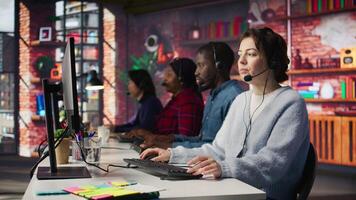 Teamworking call center coworkers in office helping customers by answering questions. Team of agents in customer support workplace in front of PC using headset mic to talk with clients, camera B video