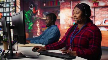 Upbeat call center agent provides guidance to customers, answering questions. Jolly African american customer center worker at PC desk walking clients through technical solutions during call, camera B video