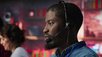 Smiling call center agent dealing with unhappy customers, speaking with them using audio headset at office desk. Customer support worker handling clients complaints in company workspace, camera A video