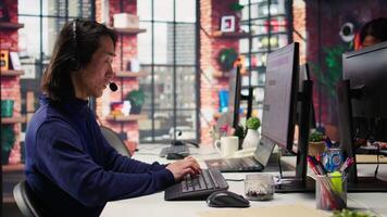 Senior call center agent helping customers regain access to accounts, resetting passwords. Experienced Asian worker in diverse customer center assisting clients needing help, camera B close up video