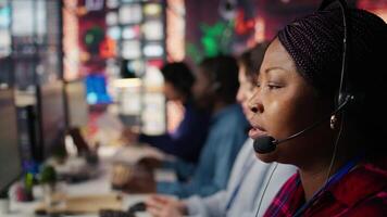 Call center employee talks in headphones mic, handling customer inquiries and complaints in office. African american customer service representative assisting customers with issues, camera B close up video