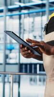 Vertical Smart factory expert using digital device app to test automated systems and ensure reliability. Worker uses tablet to identify bottlenecks and optimize gear based on sensors data, camera A close up video