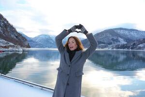 Cheerful woman making heart gesture by snowy fjord in Norway winter scene photo